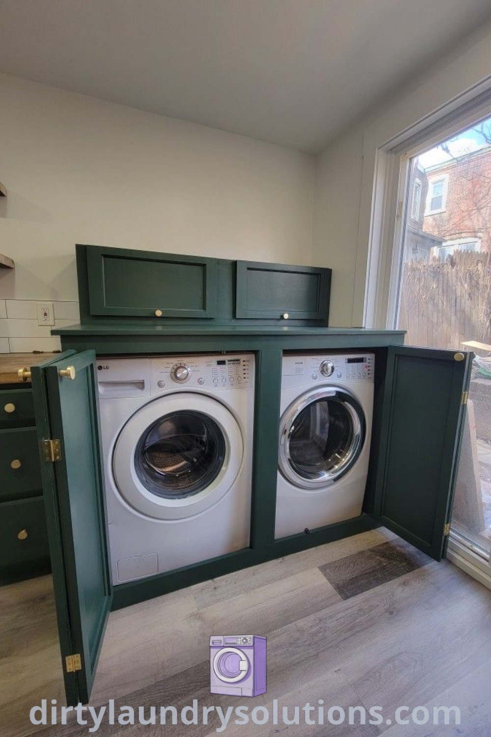 A washer and dryer in a kitchen next to an open window with green cabinets, offering a stylish hidden washer and dryer solution. Explore laundry room cabinet ideas and more at dirtylaundrysolutions.com.