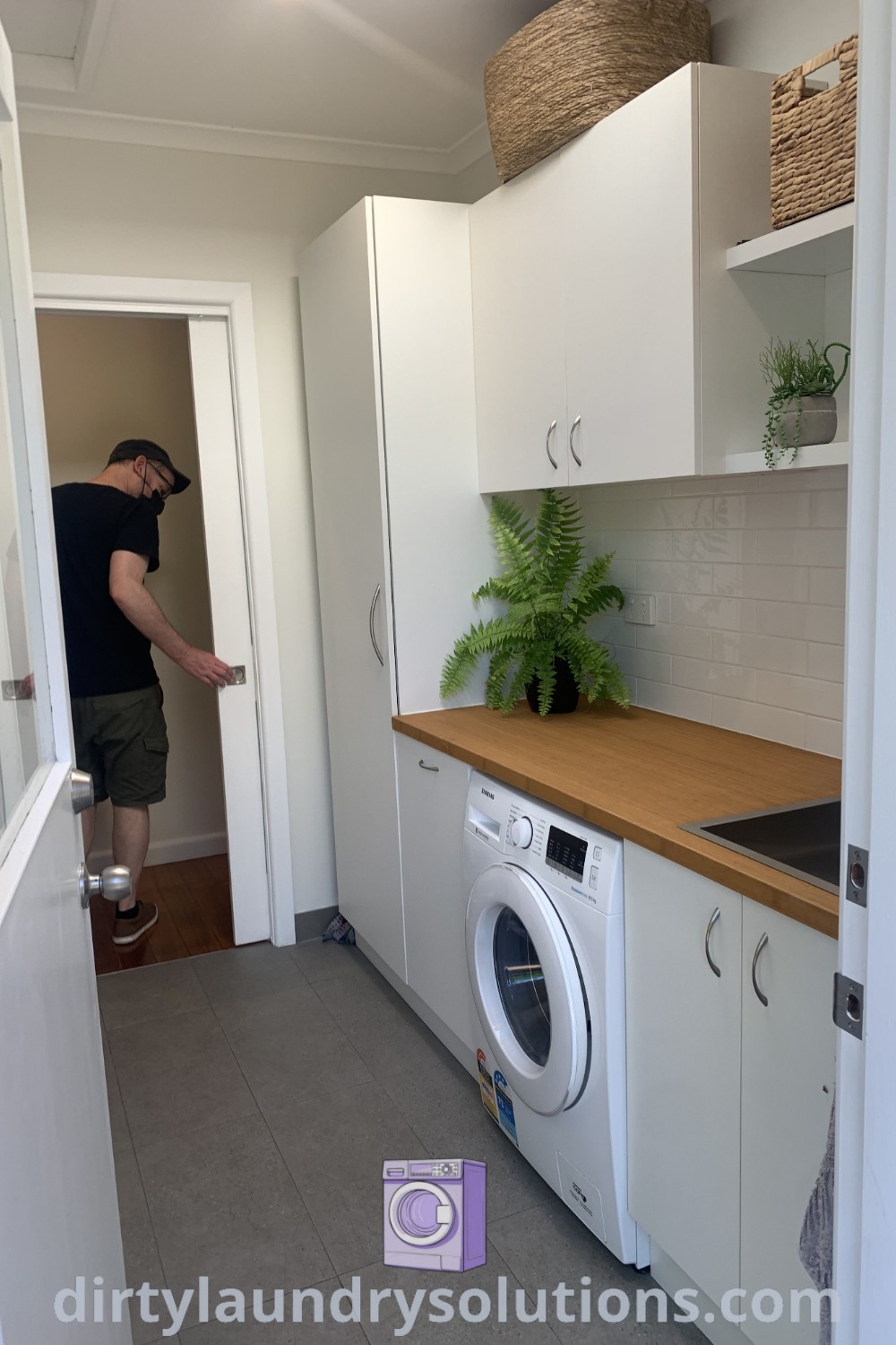 A man standing in the doorway of a cozy utility room featuring a laundry cupboard with sink, raised washing machine, and dryer, showcasing stylish laundry room ideas in a compact design. Discover more unique ideas and solutions for your home at dirtylaundrysolutions.com.