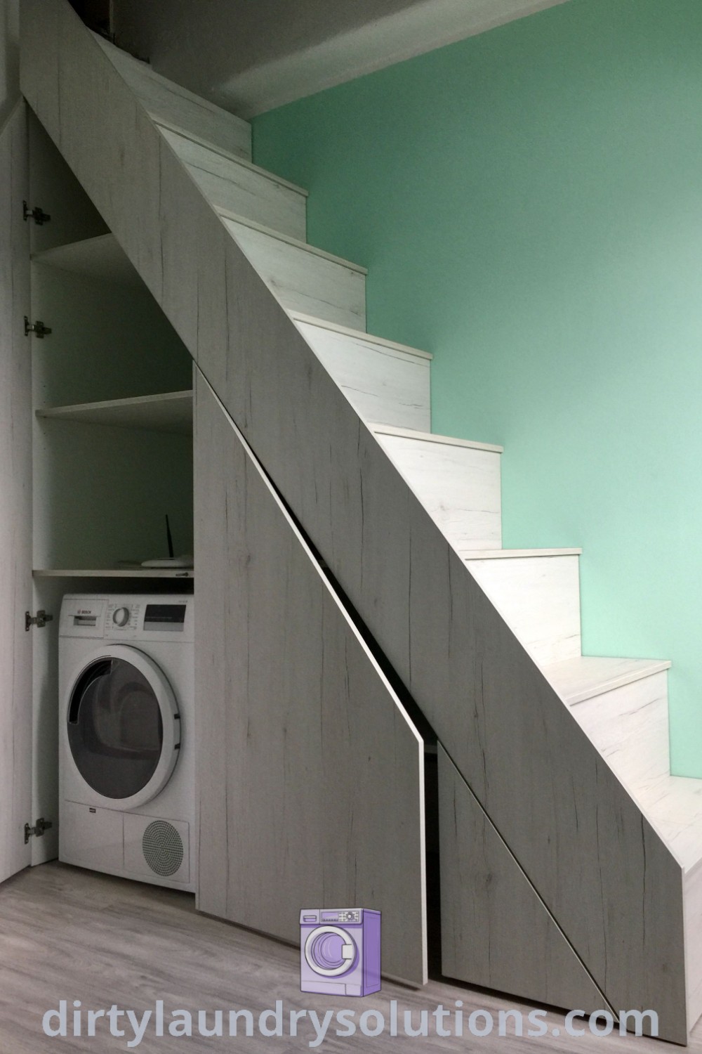 A washer and dryer under a staircase in a room with green walls, showcasing effective use of space and inspiring design ideas for small homes. For more solutions to organize your laundry area and enhance your cozy home aesthetic, visit dirtylaundrysolutions.com.